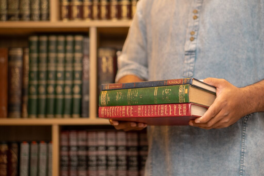 A man wearing a denim shirt holds stacked Arabic books in a library, symbolizing study and learning.